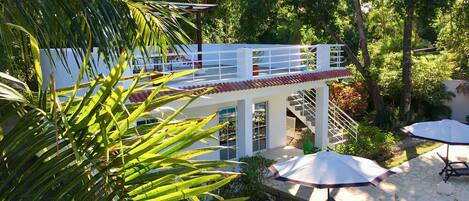 A two-story building with a white exterior and red roof, surrounded by lush greenery and a swimming pool area with umbrellas and lounge chairs.