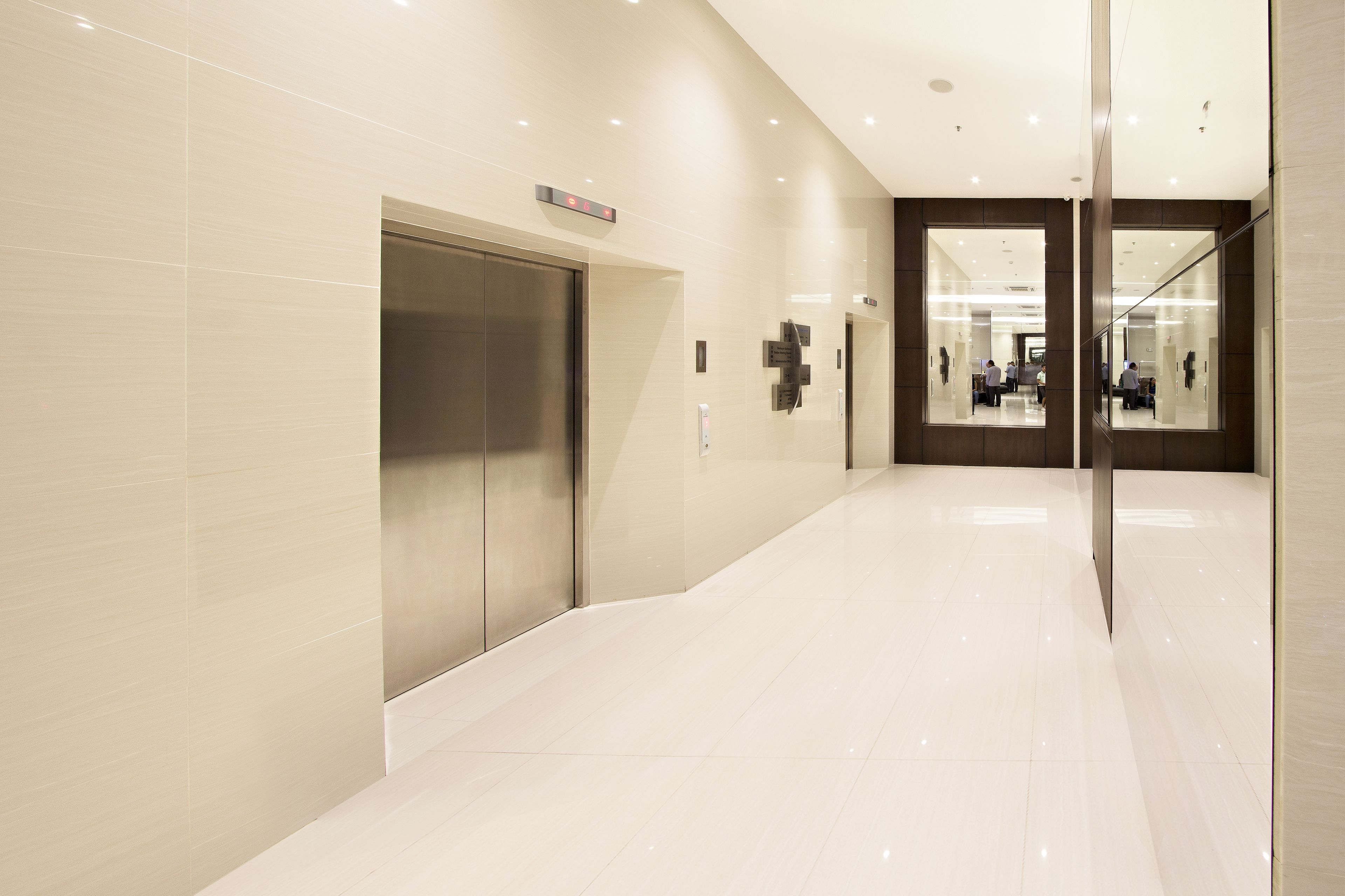 A modern, well-lit hallway with a shiny floor, a digital elevator display, and a glass door leading to a room with a view.