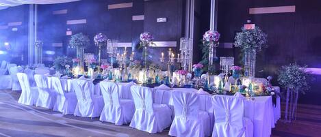 A banquet hall with tables set for a formal event, featuring white tablecloths, floral centerpieces, and candlelit arrangements.