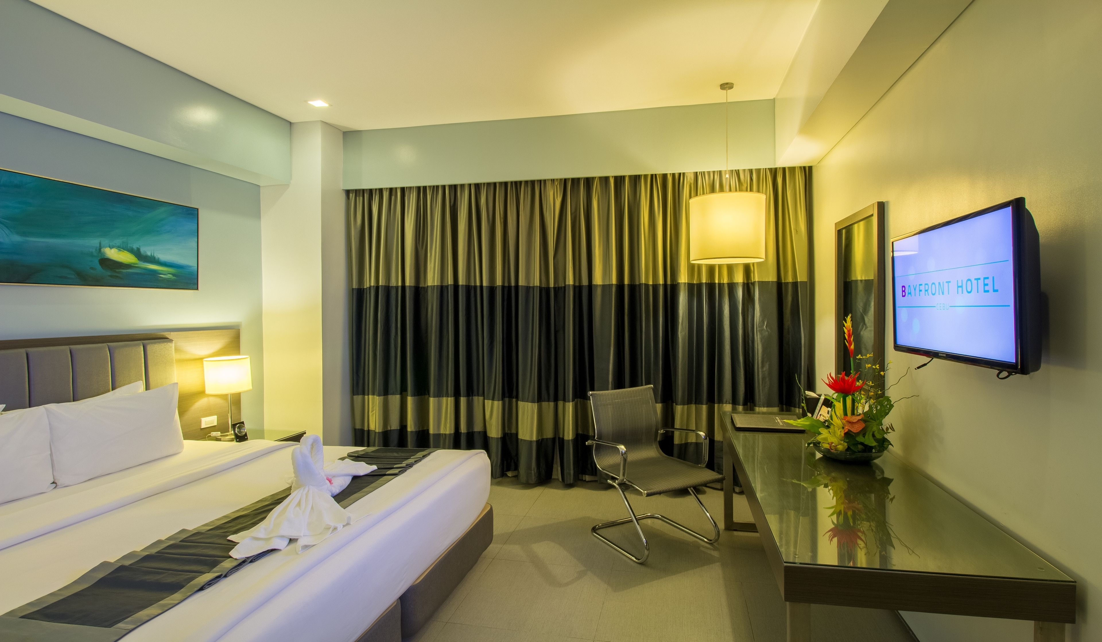 A hotel room with a bed, a desk, a chair, a TV, and a painting on the wall.