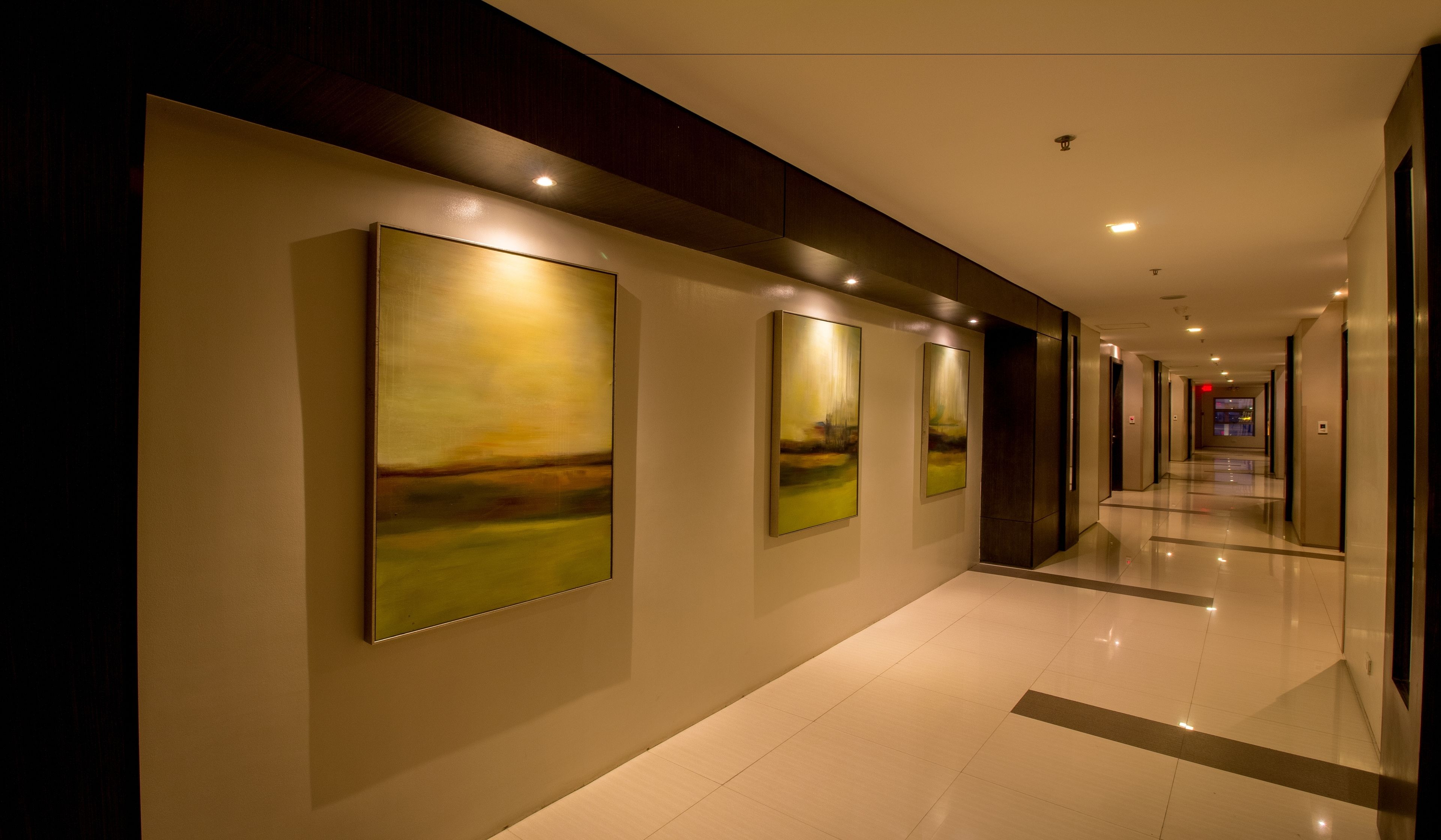 A modern hotel corridor with artwork on the walls, a clean tiled floor, and a long hallway leading to an elevator.