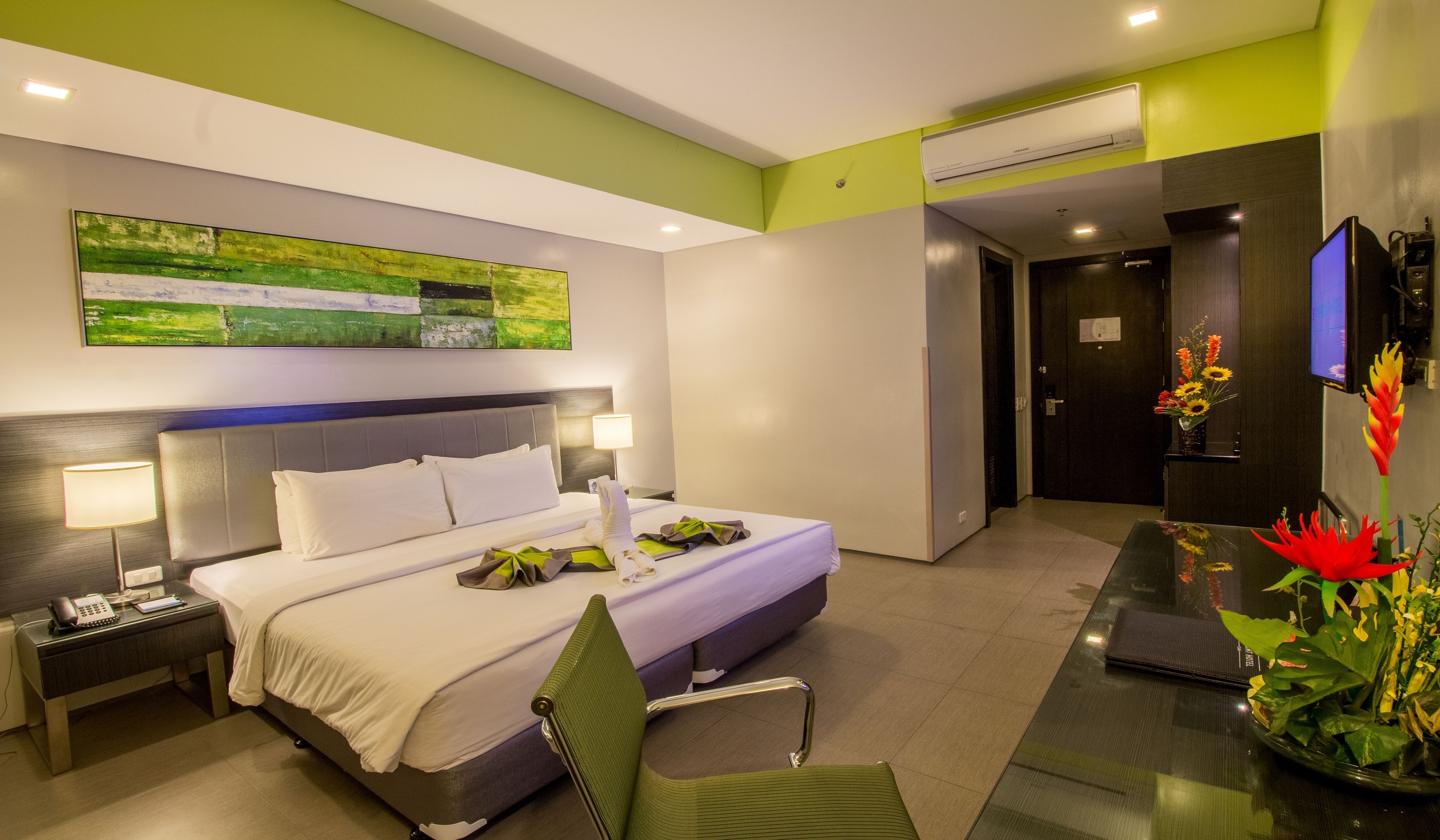 Una habitación de hotel moderna con una cama grande, mesitas de noche con lámparas, un televisor de pantalla plana, un escritorio con una silla y una pared verde con un cuadro.