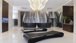 A modern hotel lobby with a reception desk, a large screen displaying news, and a lounge area with black sofas.
