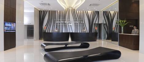 A modern hotel lobby with a reception desk, a large screen displaying news, and a lounge area with black sofas.