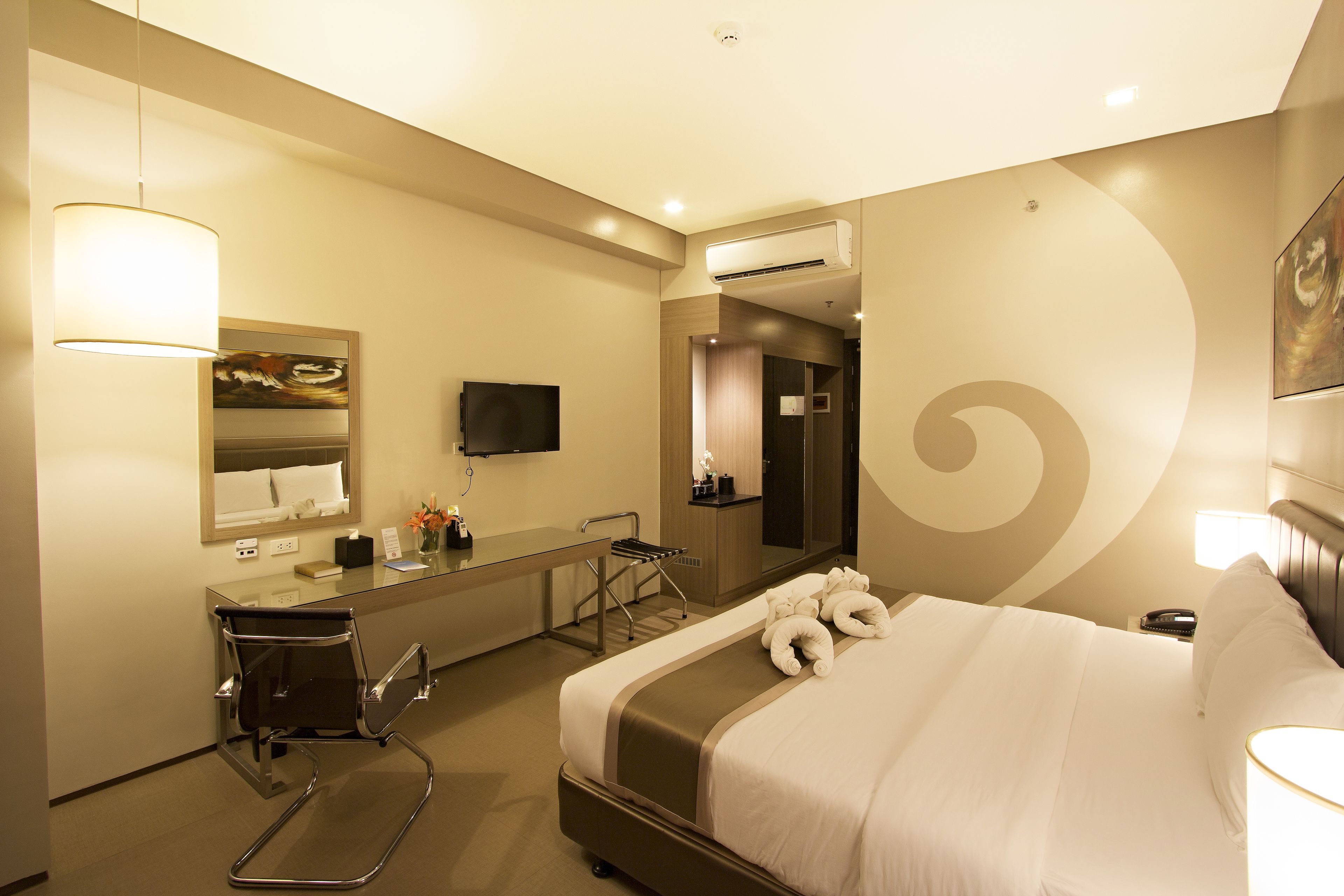 A hotel room with a bed, a desk with a chair, a mirror, a television, and an air conditioning unit.