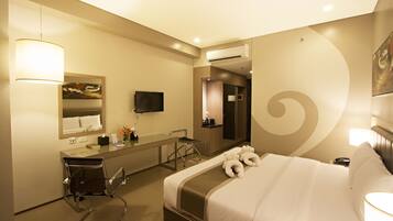 A hotel room with a bed, a desk with a chair, a mirror, a television, and an air conditioning unit.