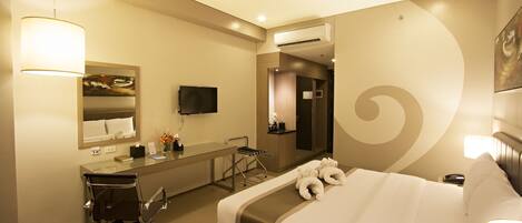 A hotel room with a bed, a desk with a chair, a mirror, a television, and an air conditioning unit.