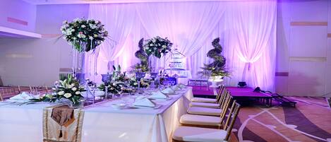 A banquet hall with a long table set for a formal event, adorned with floral centerpieces and elegant tableware.