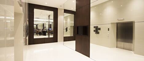 A modern interior with a glass wall reflecting a busy area, a mounted TV, and an elevator.