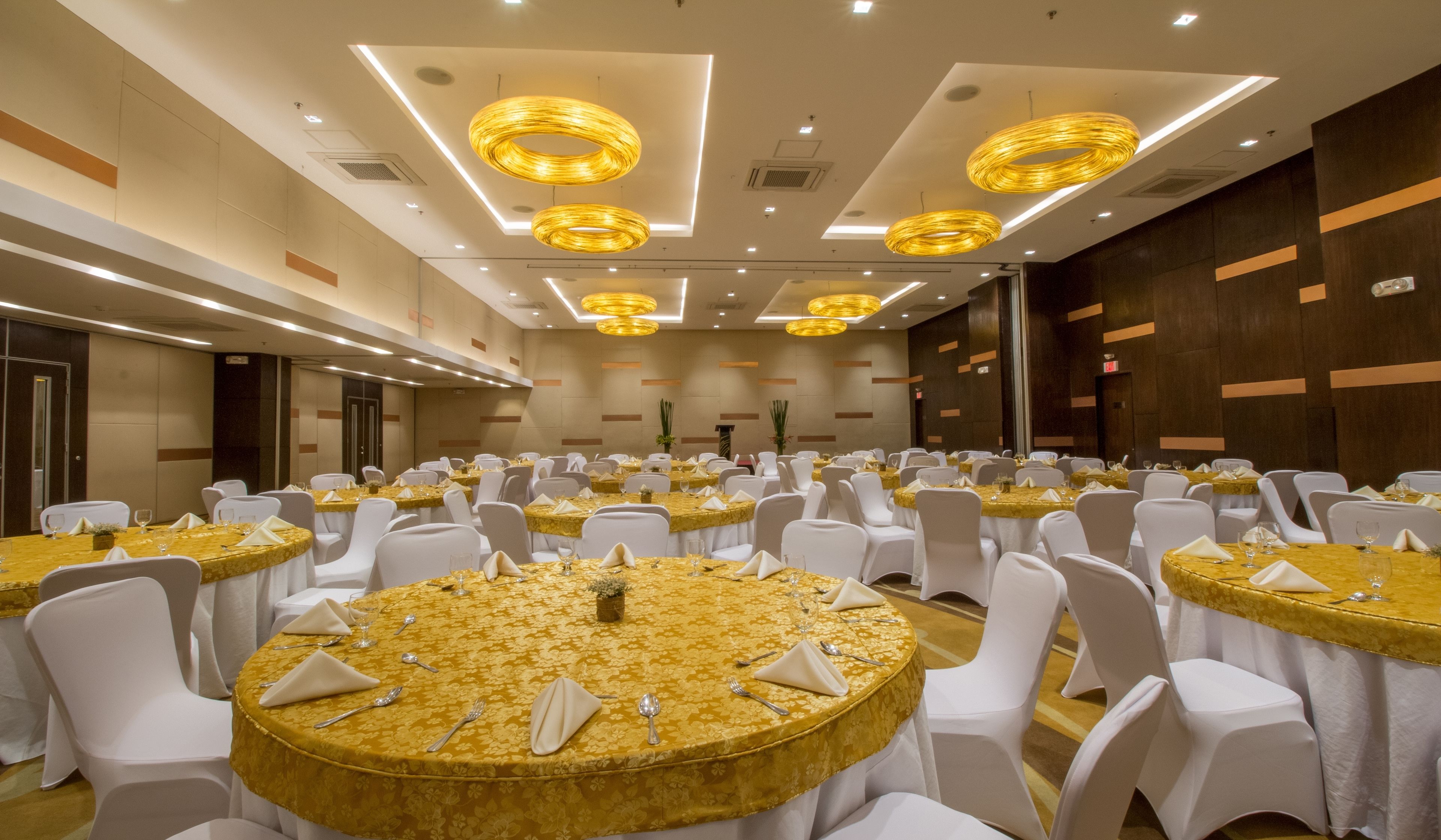 Une salle de banquet avec des tables rondes recouvertes de nappes dorées, des chaises blanches et des luminaires modernes.