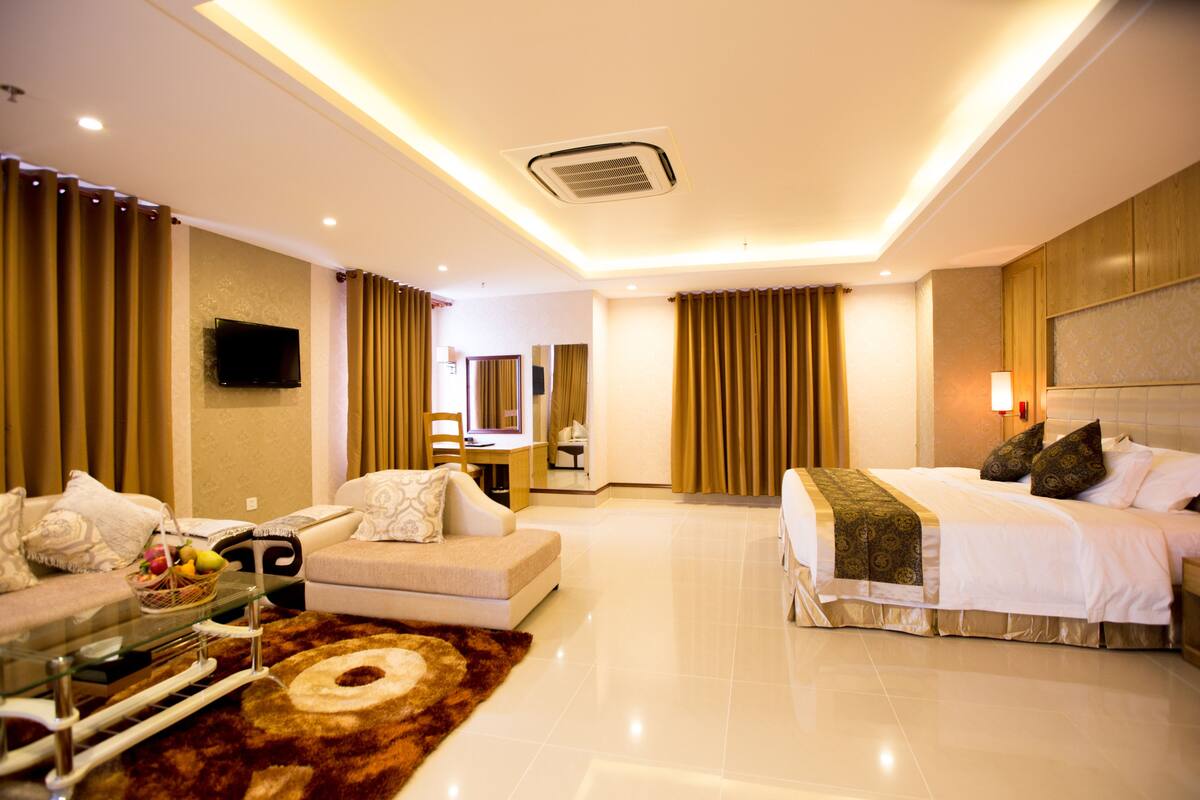 A modern hotel room with a large bed, a sofa, a TV, and a dining area.