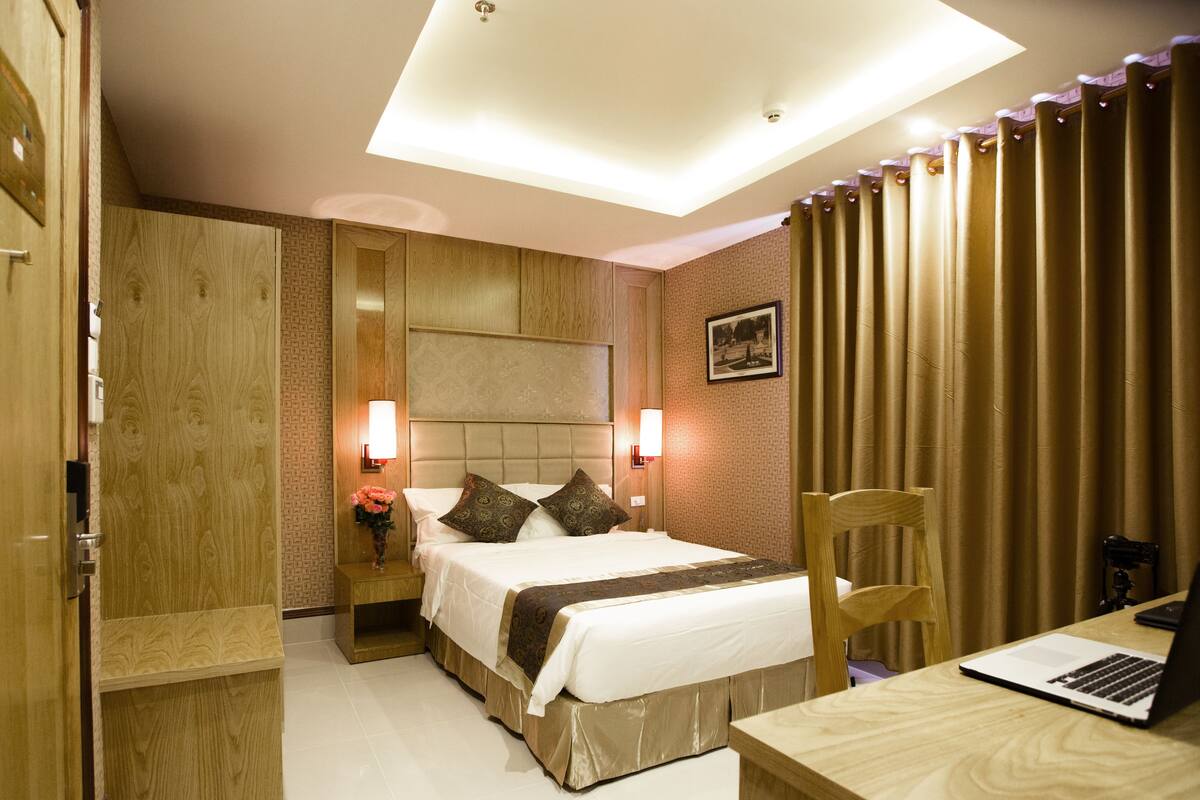 A hotel room with a bed, a desk with a laptop, a chair, a nightstand with a vase, and a wall-mounted headboard.