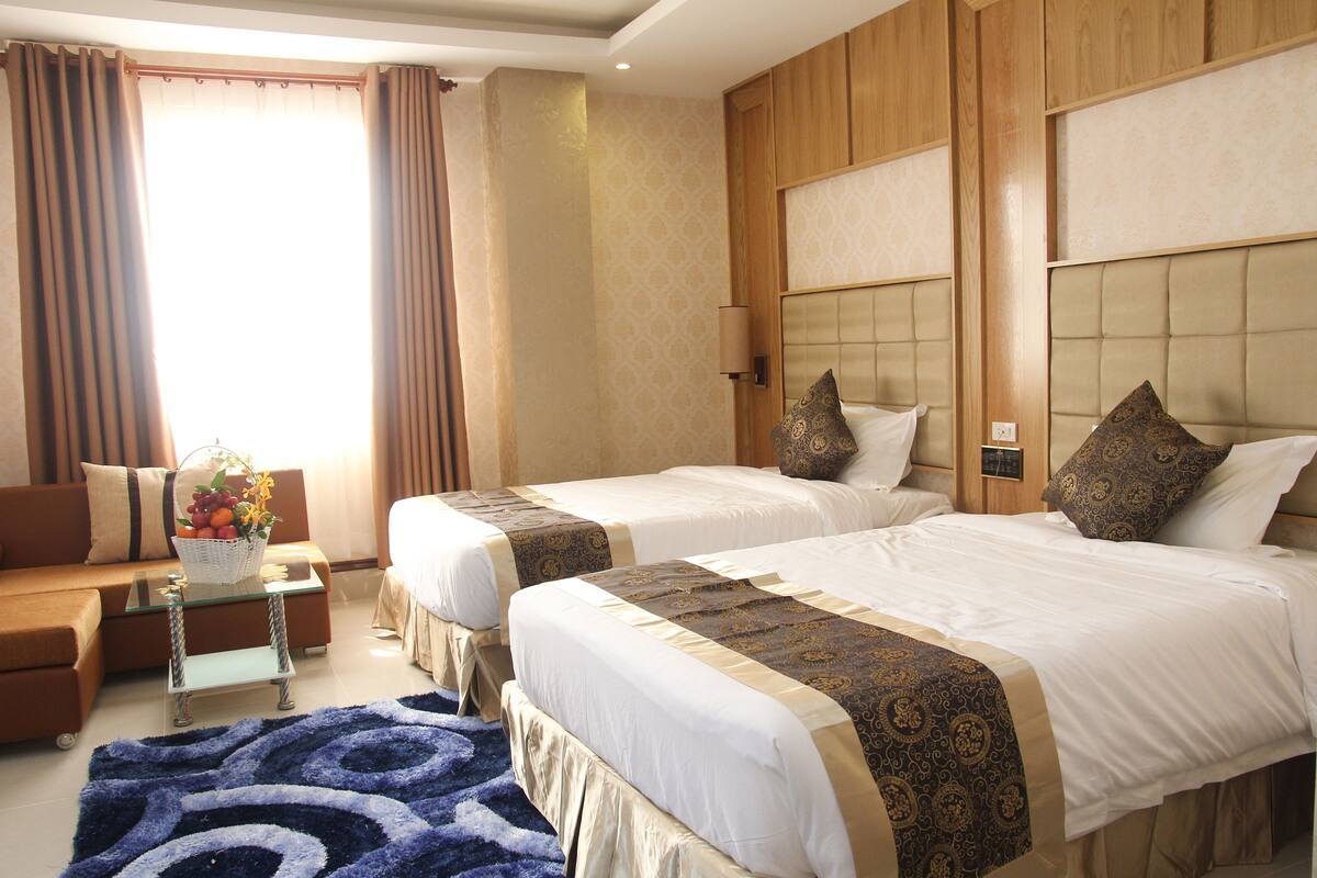 A hotel room with two beds, a sofa, a small table with a fruit basket, and a patterned rug.