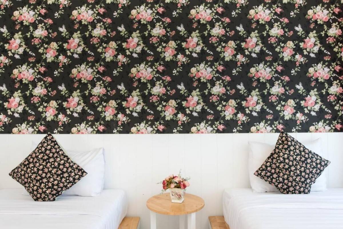 Een kamer met bloemenbehang, twee witte bedden met bijpassende kussens en een klein rond houten tafeltje met een bloevas.