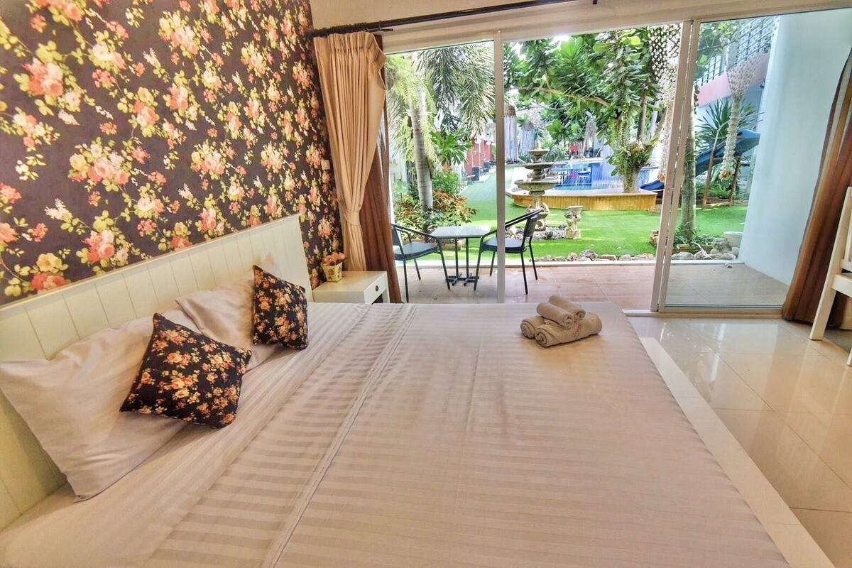 Een slaapkamer met een bed, kussens, een klein tafeltje en een stoel. Er is uitzicht op een tuin door een glazen deur.
