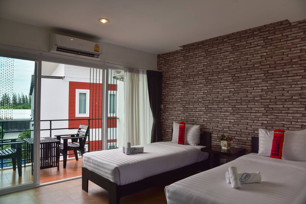 Hotelkamer met twee bedden, een bakstenen muur, een balkon met een tafel en stoelen, en uitzicht op een gebouw met een rood-witte gevel.