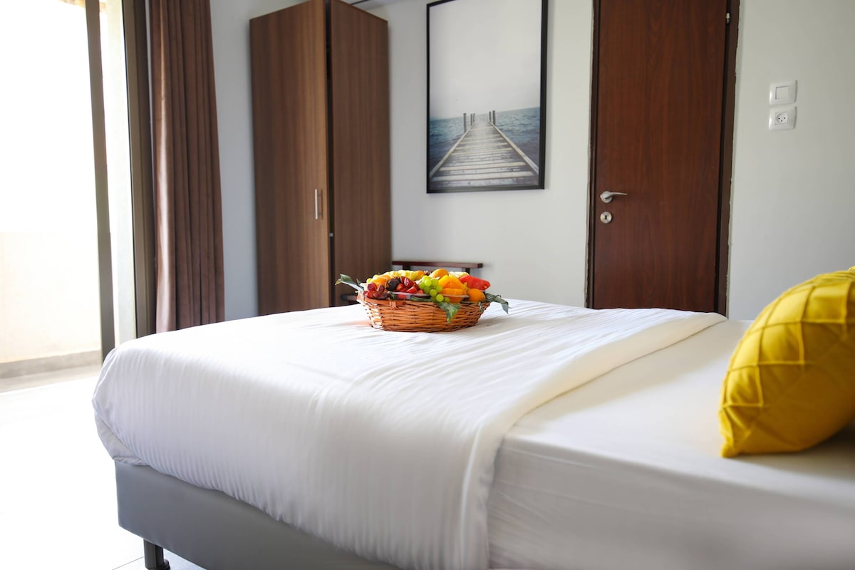 A neatly made bed with a white comforter and a yellow pillow, a fruit basket on the bed, and a wooden wardrobe with a picture of a pier on the wall.