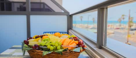 Una cesta de mimbre con fruta sobre una mesa junto a una ventana con vistas a la playa.