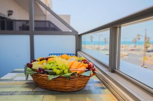 Una cesta de mimbre con fruta sobre una mesa junto a una ventana con vistas a la playa.