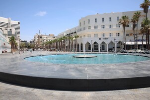 Una plaza pública con una piscina central, rodeada de palmeras y edificios con comercios.