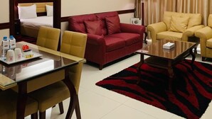 A hotel room with a bed, a red sofa, a yellow sofa, a dining table with chairs, a coffee table, a lamp, and a red and black rug. - Hala Inn Hotel Apartments (Ajman)