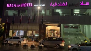 The exterior of the Hala Inn Hotel with a prominent neon sign, multiple flags, and a parking area with several cars. - Hala Inn Hotel Apartments (Ajman)