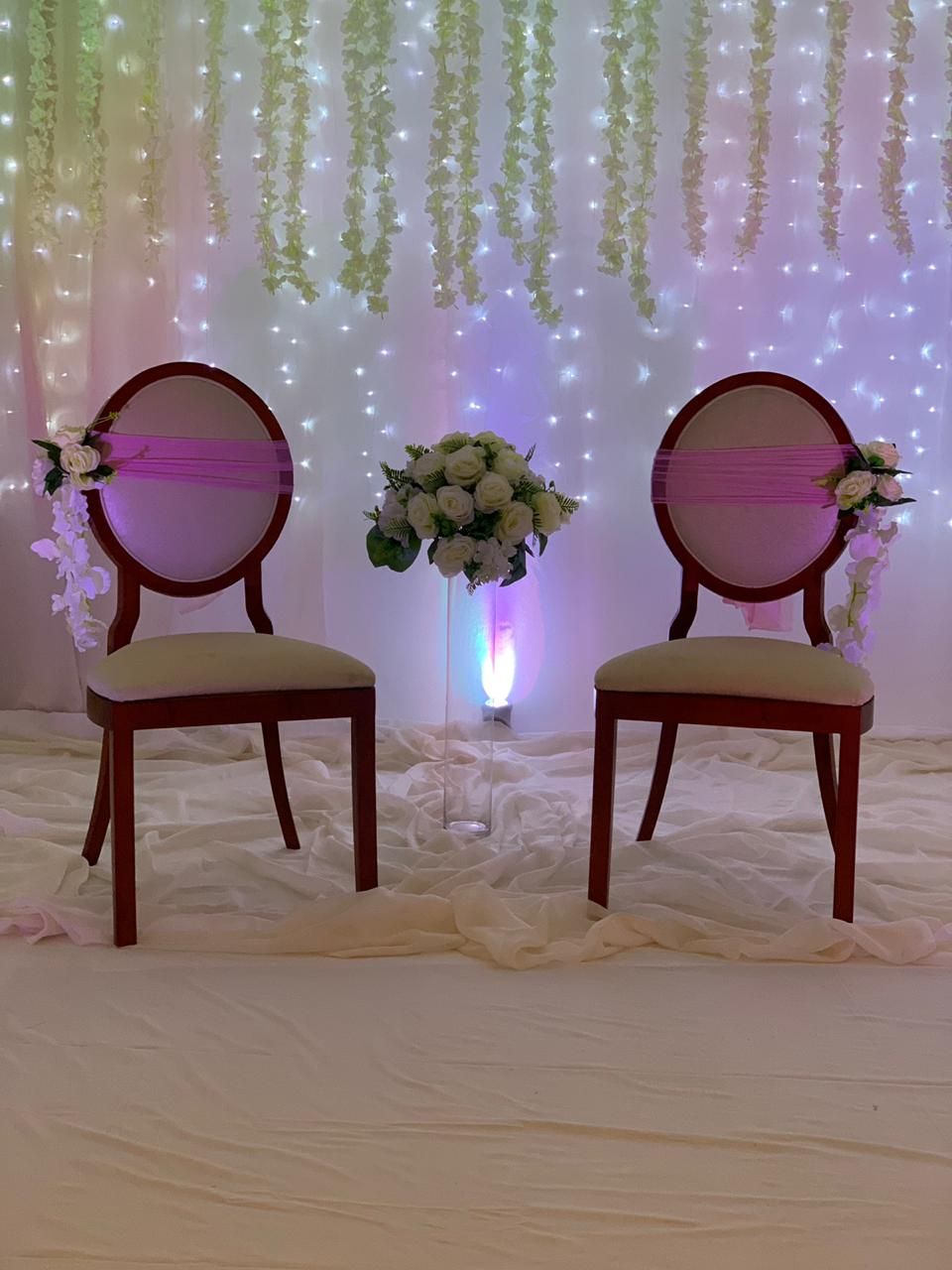 Two wooden chairs with cushioned seats and backs, positioned on a white fabric-covered floor, with a floral arrangement between them.