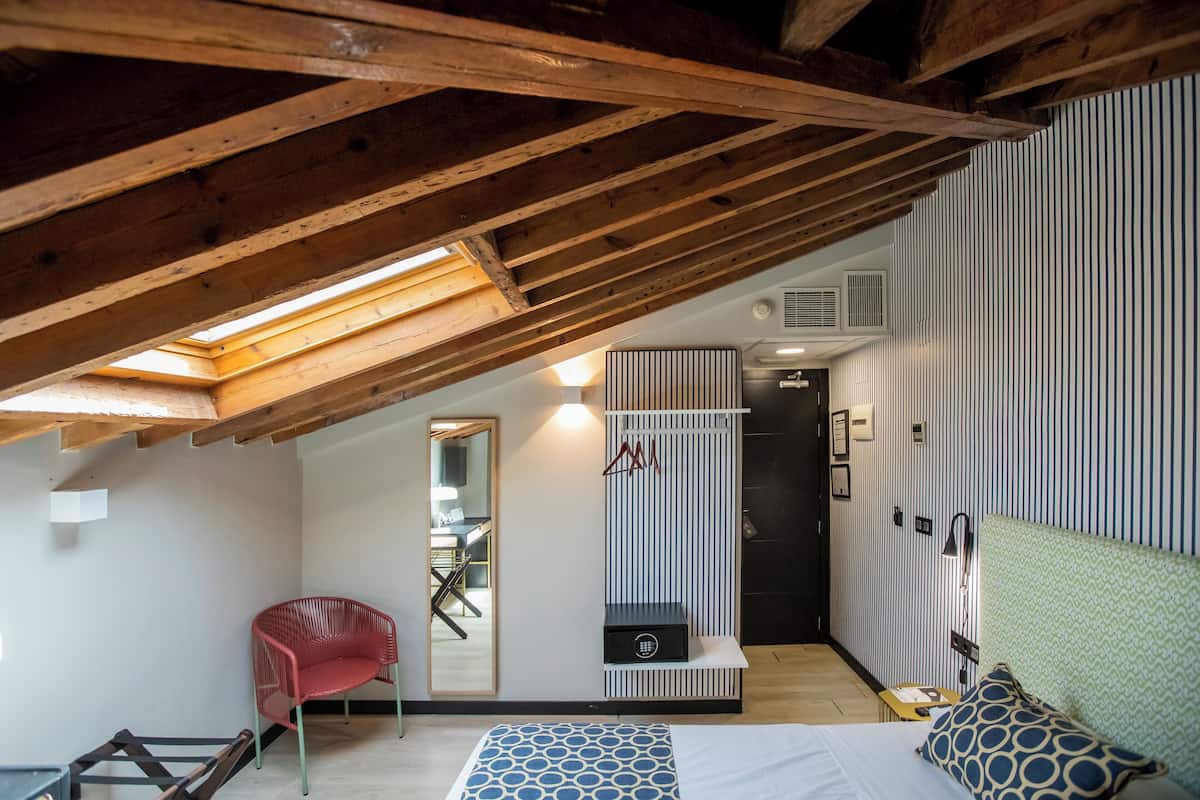 A bedroom with a sloped ceiling, a bed with patterned bedding, a red chair, a mirror, and a wardrobe.