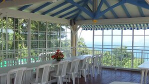 A covered outdoor dining area with white plastic chairs and tables, large windows, and a view of the ocean. - Résidence Bel'Bay (Les Anses-d'Arlets)