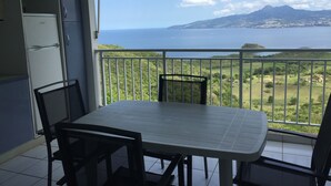 A balcony with a table and chairs, overlooking a coastal landscape with mountains and a body of water. - Résidence Bel'Bay (Les Anses-d'Arlets)