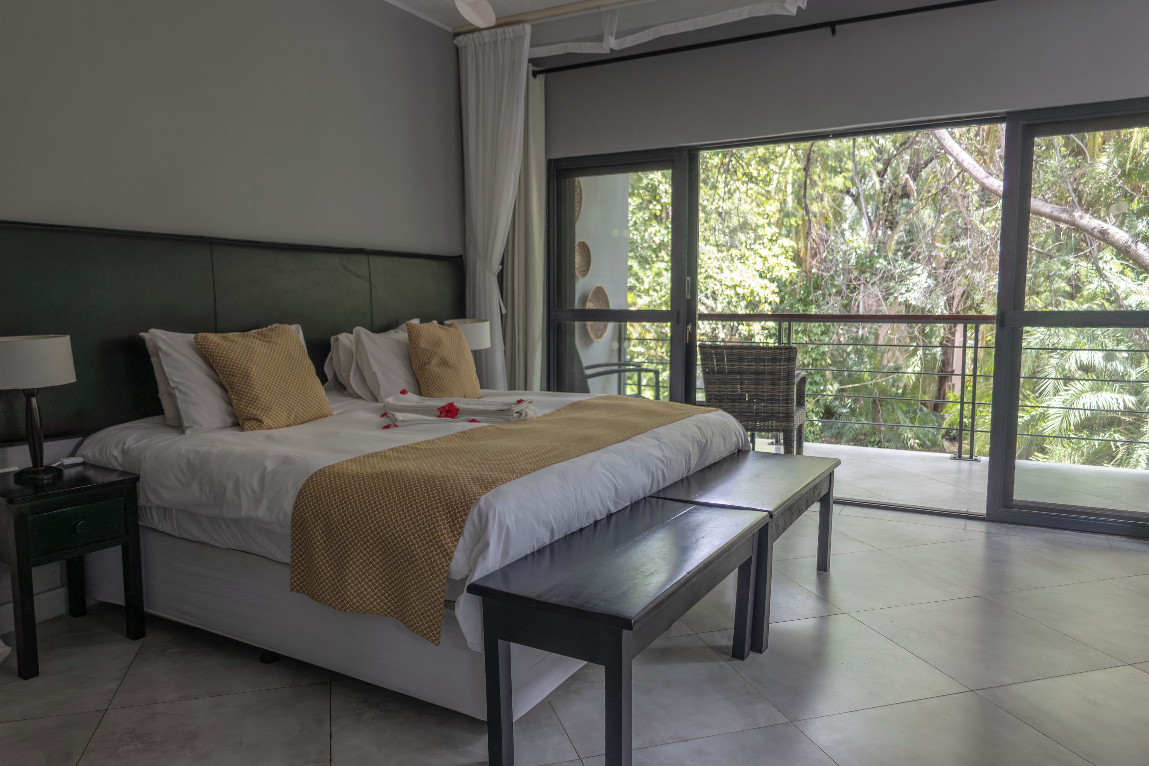 Quarto moderno com cama grande, um banquinho, mesa de cabeceira e vista para árvores por uma janela ampla.