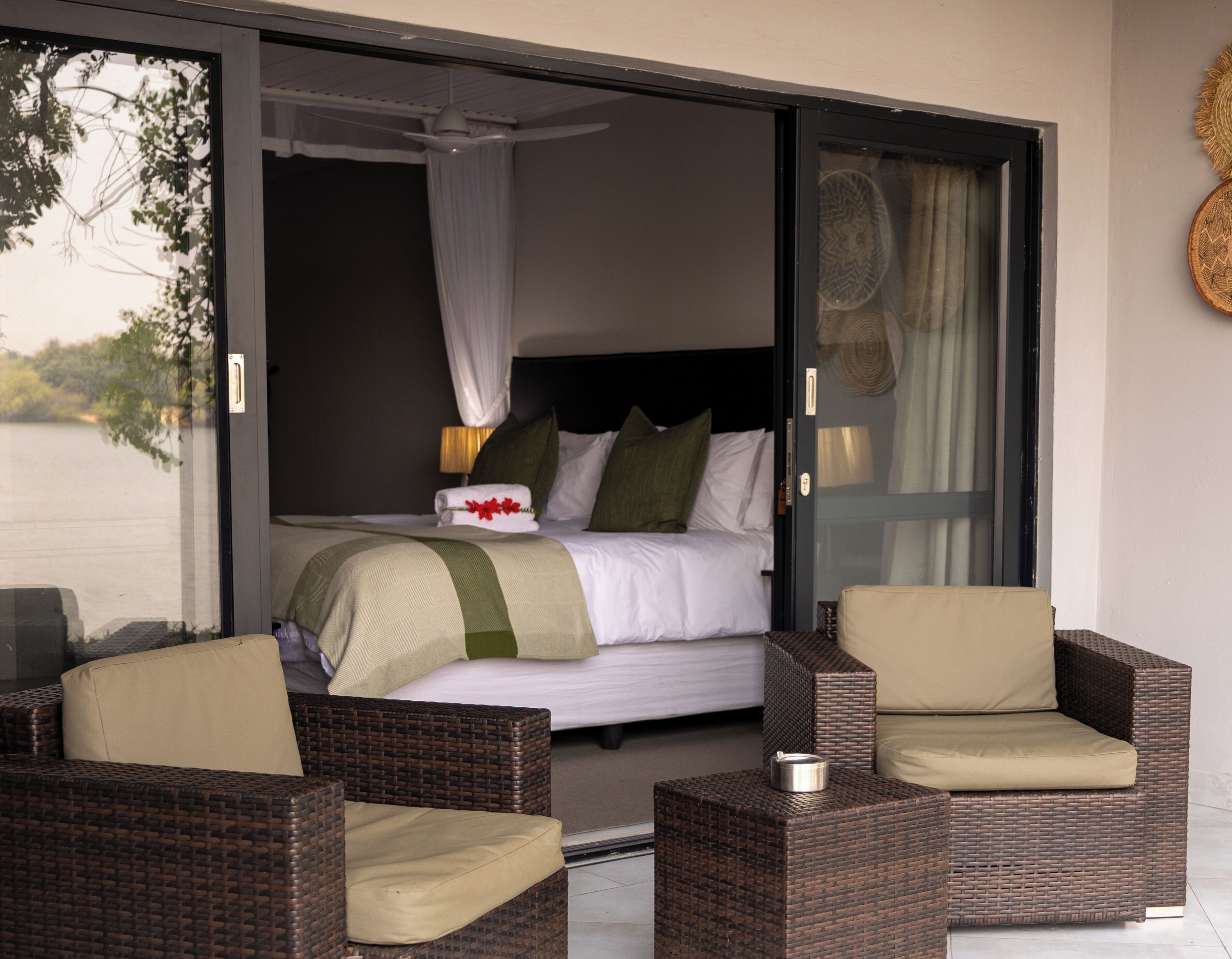 Une chambre à coucher avec un lit, un baldaquin et une vue sur un plan d’eau. On y trouve deux fauteuils en osier et une petite table avec une bougie.