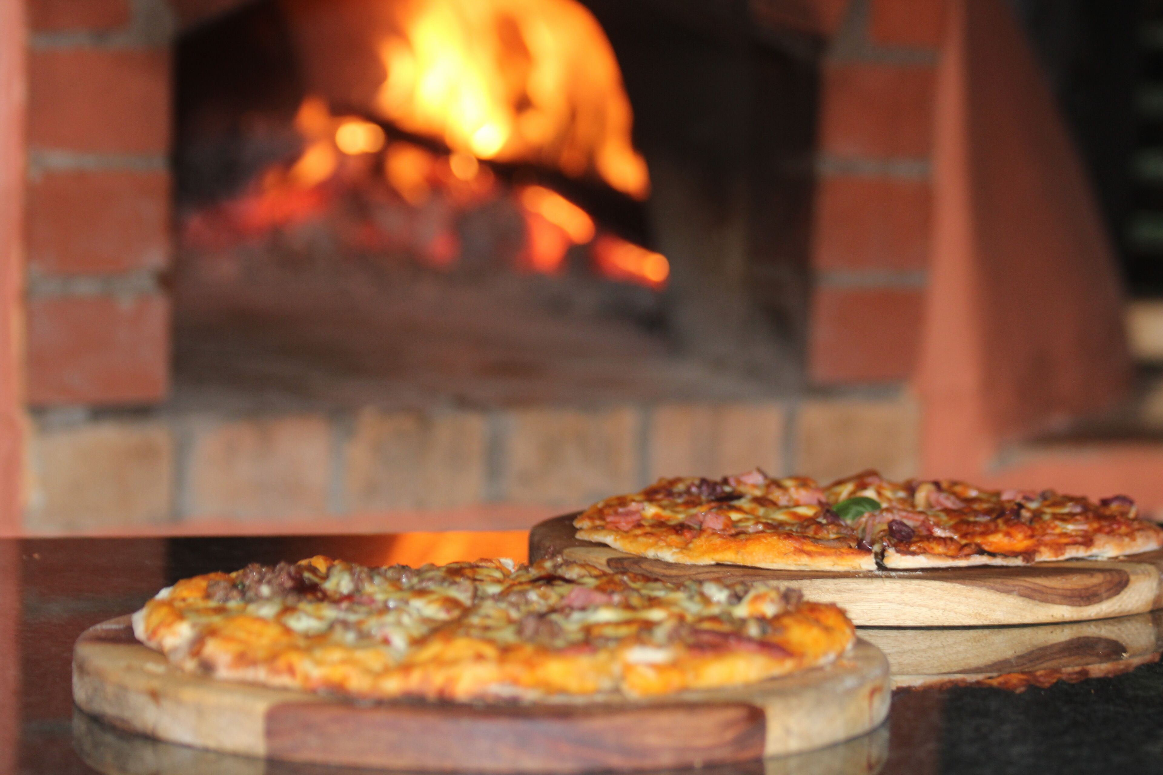 Two pizzas on wooden boards in front of a brick oven with a fire.