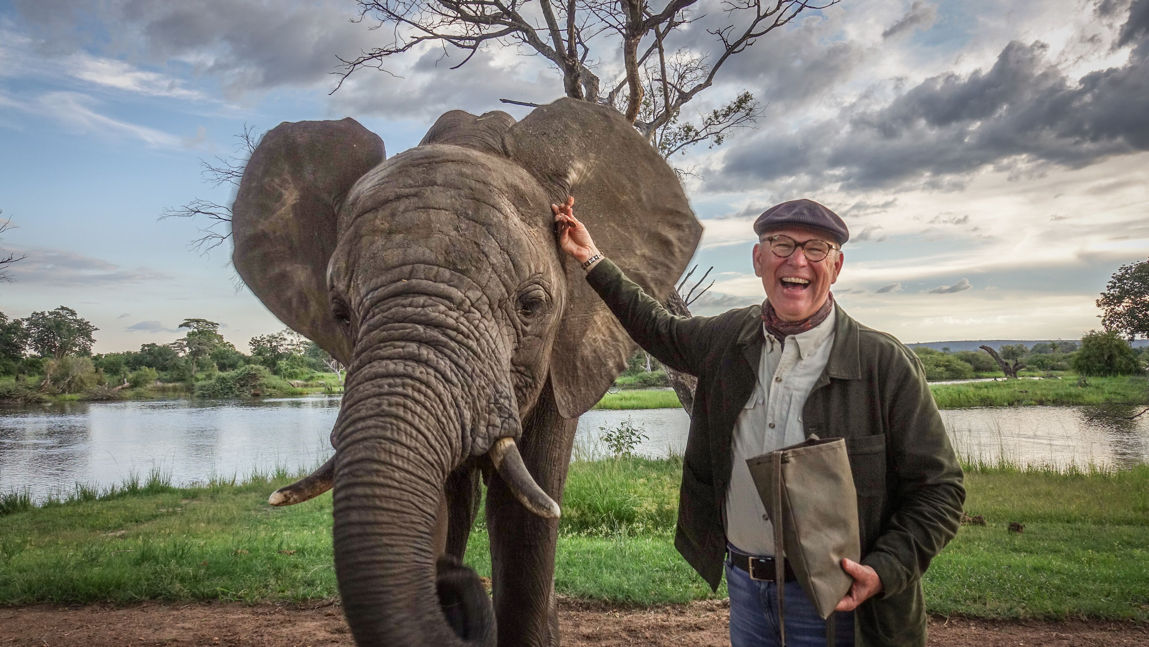 A man standing next to an elephant by a body of water with trees in the background.