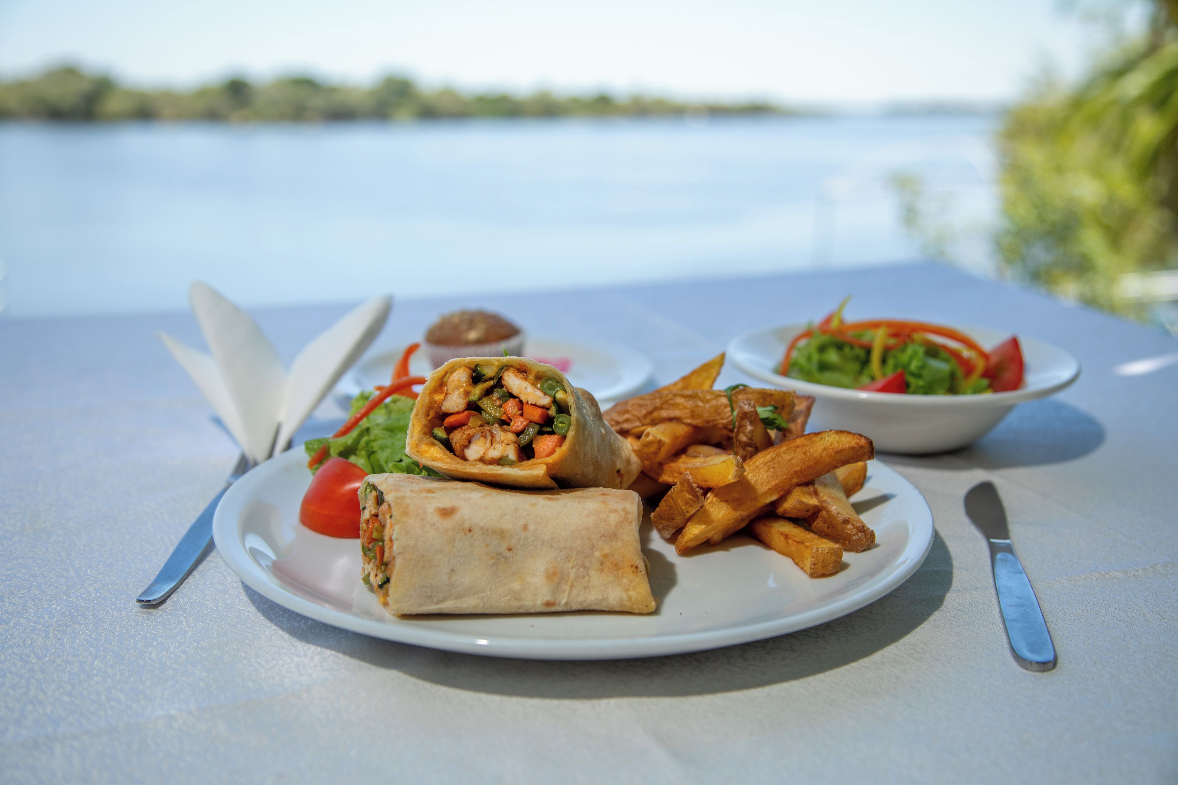 A meal consisting of a wrap, fries, and a salad on a table with a view of a body of water.