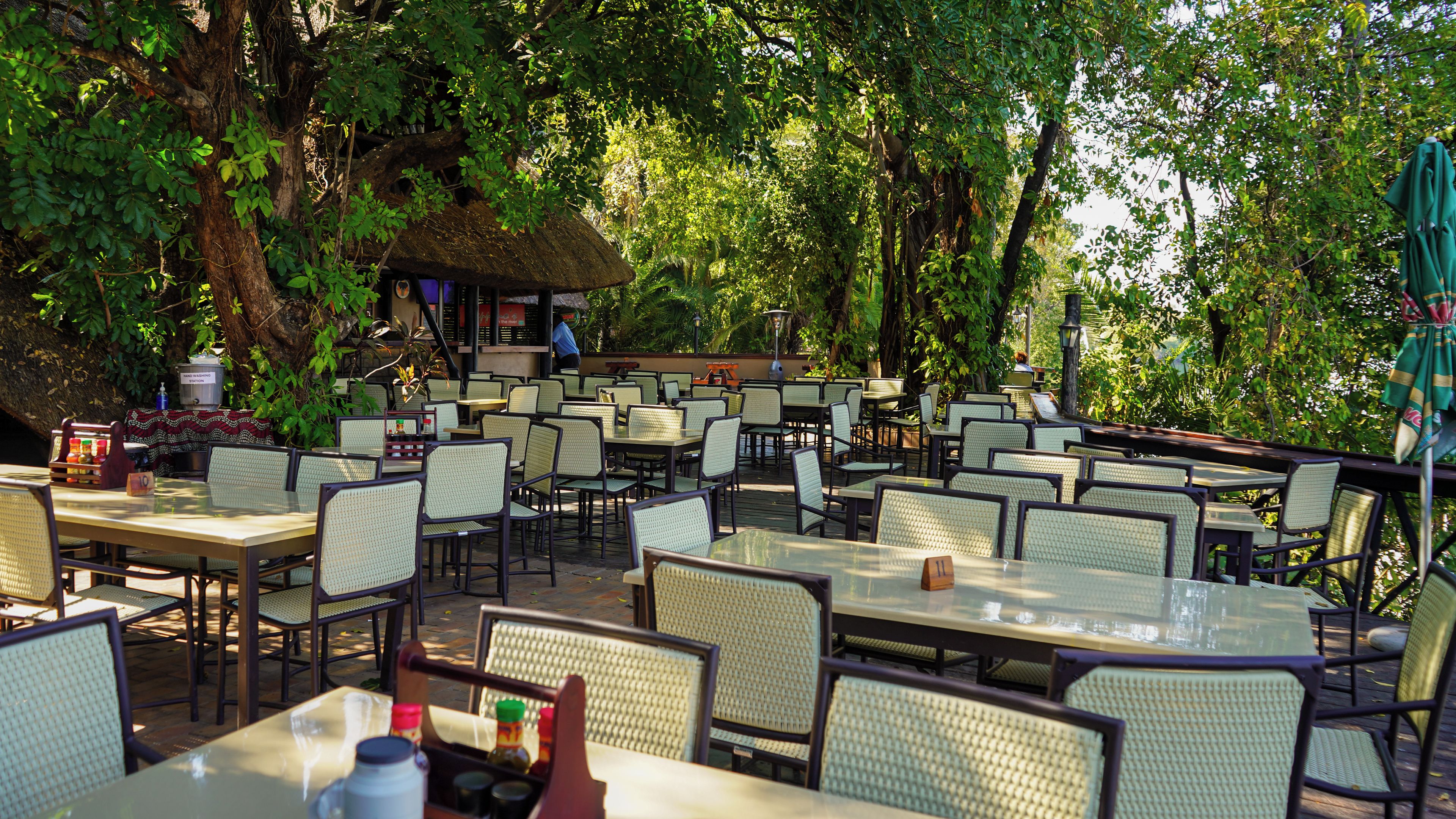 An outdoor dining area with multiple tables and chairs, surrounded by lush greenery and trees.