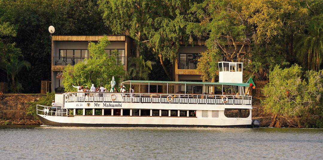 A riverboat with a deck area, passengers, and a building in the background.