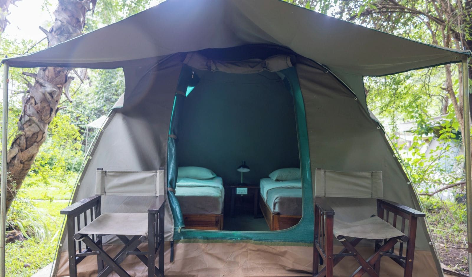 Een tent met twee bedden en twee stoelen, gelegen in een bebost gebied.