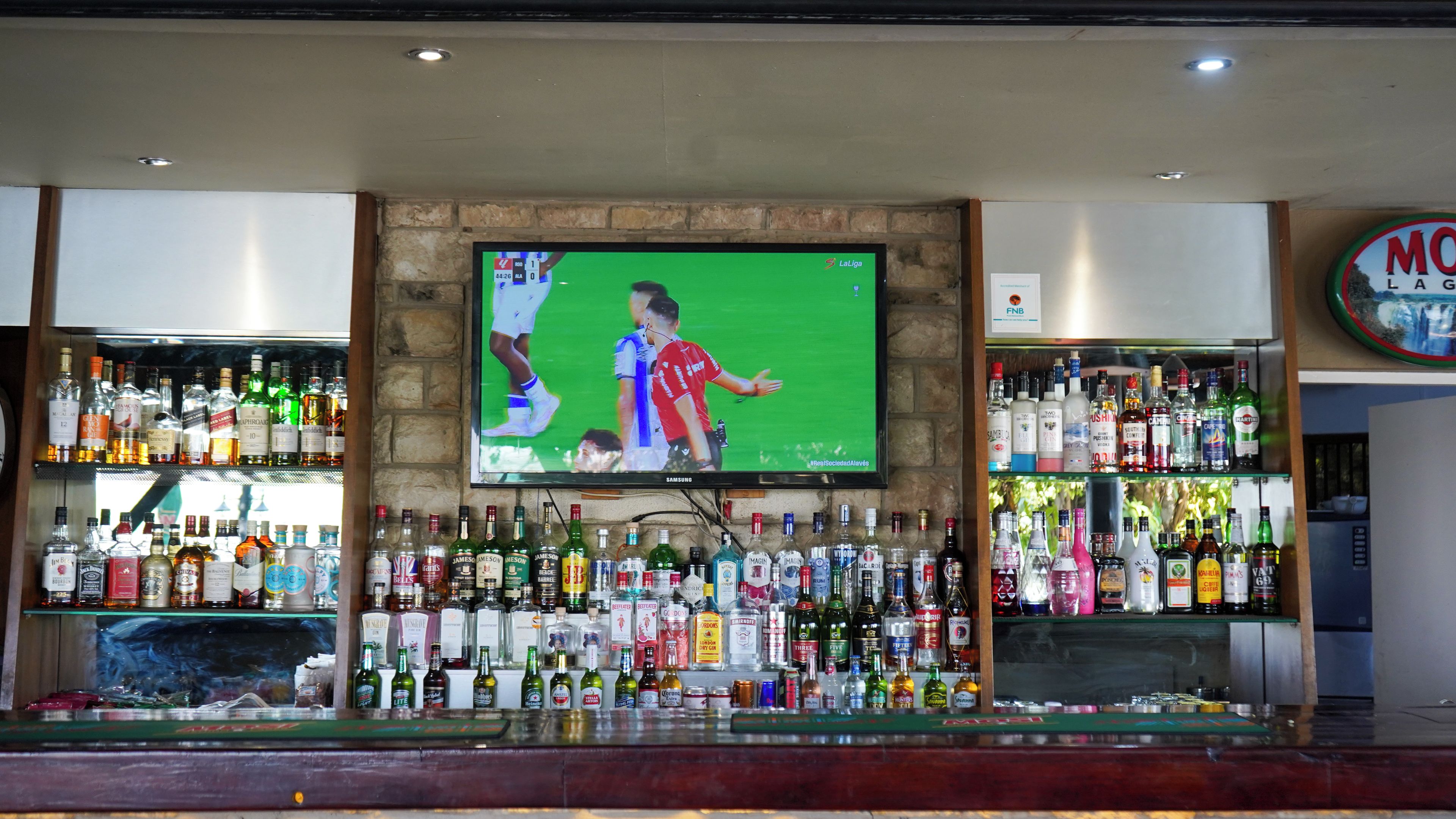 Uma área de bar com uma televisão exibindo um evento esportivo, prateleiras cheias de garrafas de bebidas alcoólicas e uma placa com o nome do bar.