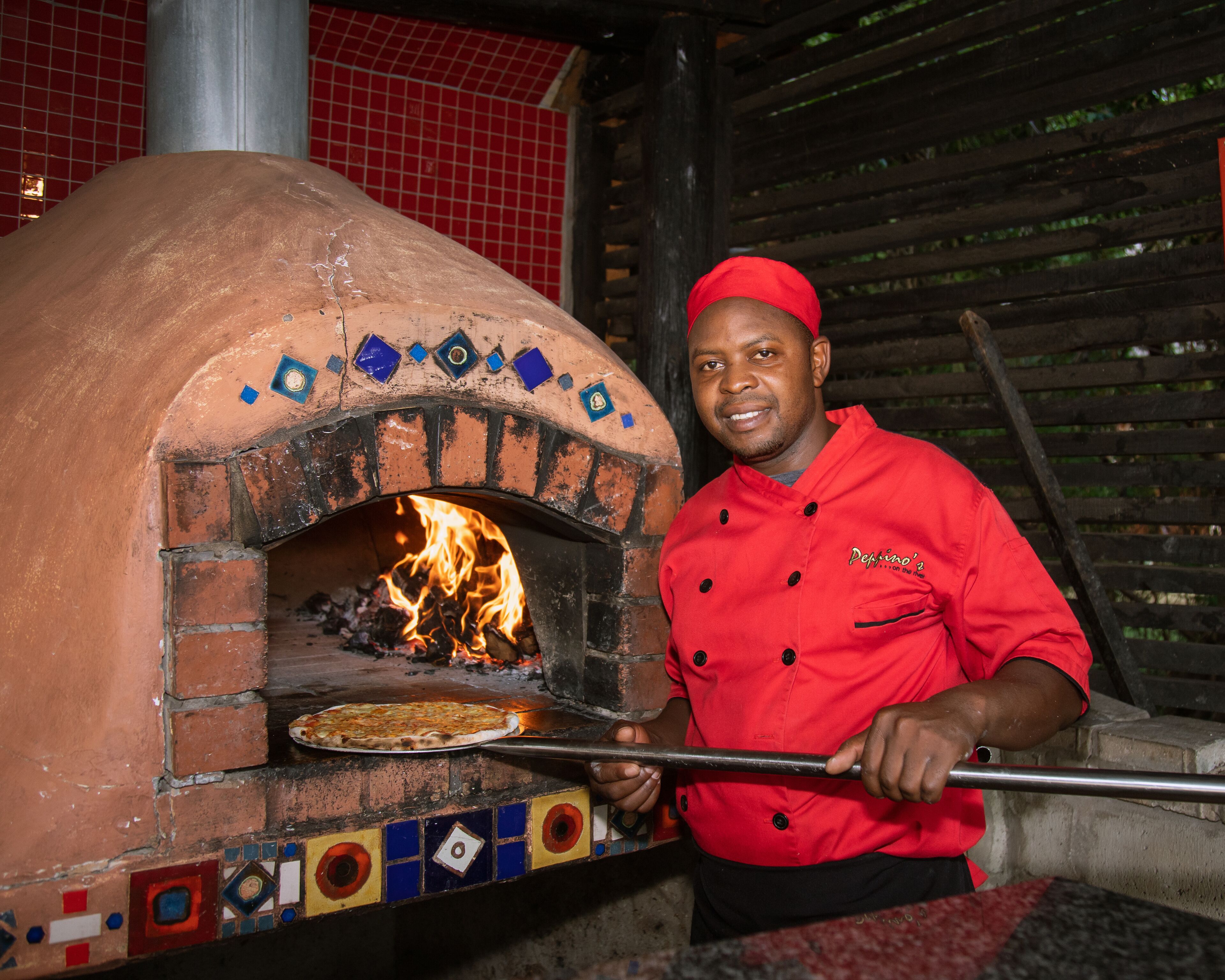 A person in a red chef's jacket operating a brick oven pizza oven.