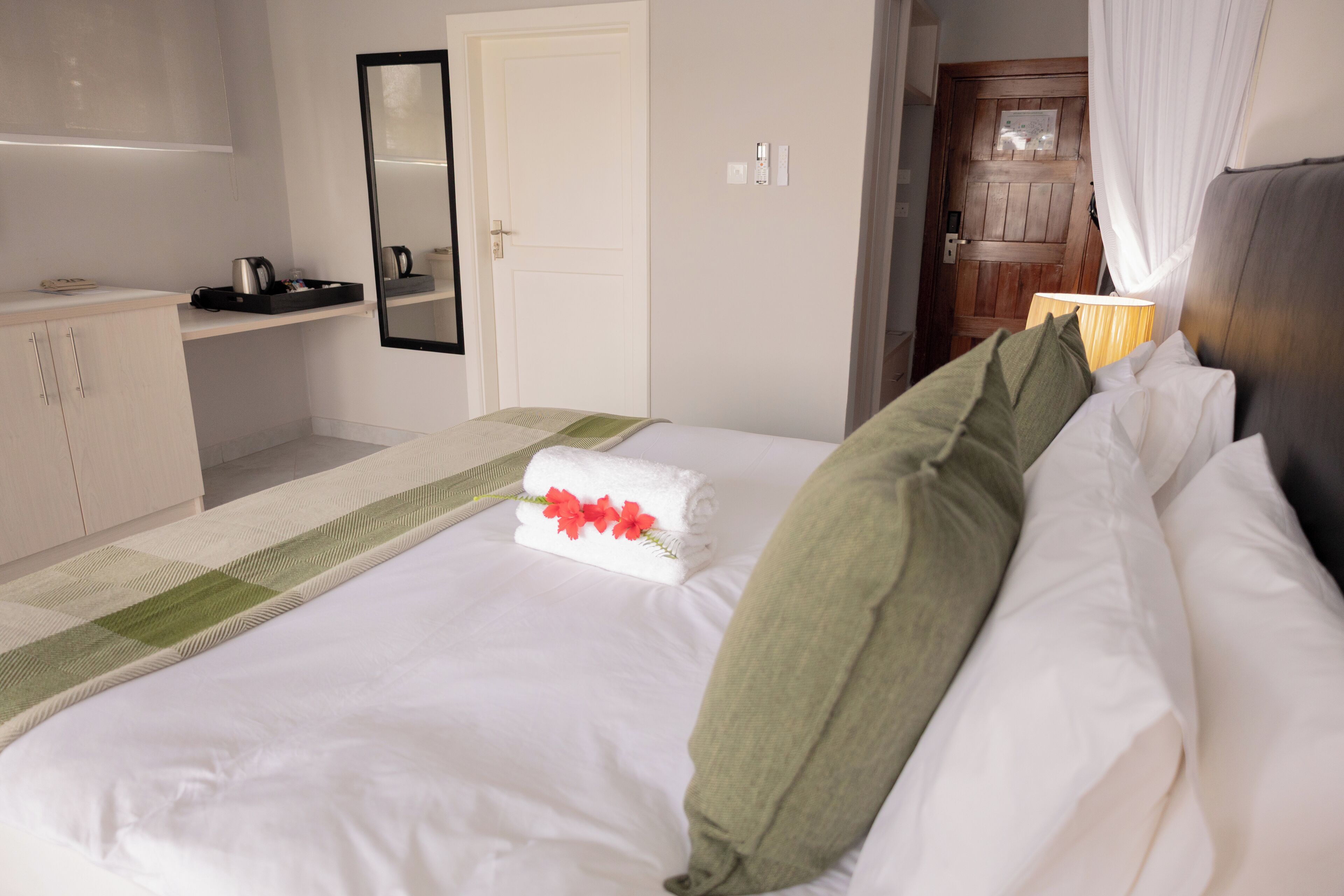 Un lit bien fait, avec du linge de lit blanc et un oreiller vert, une table de chevet avec une lampe, et une porte en bois en arrière-plan.