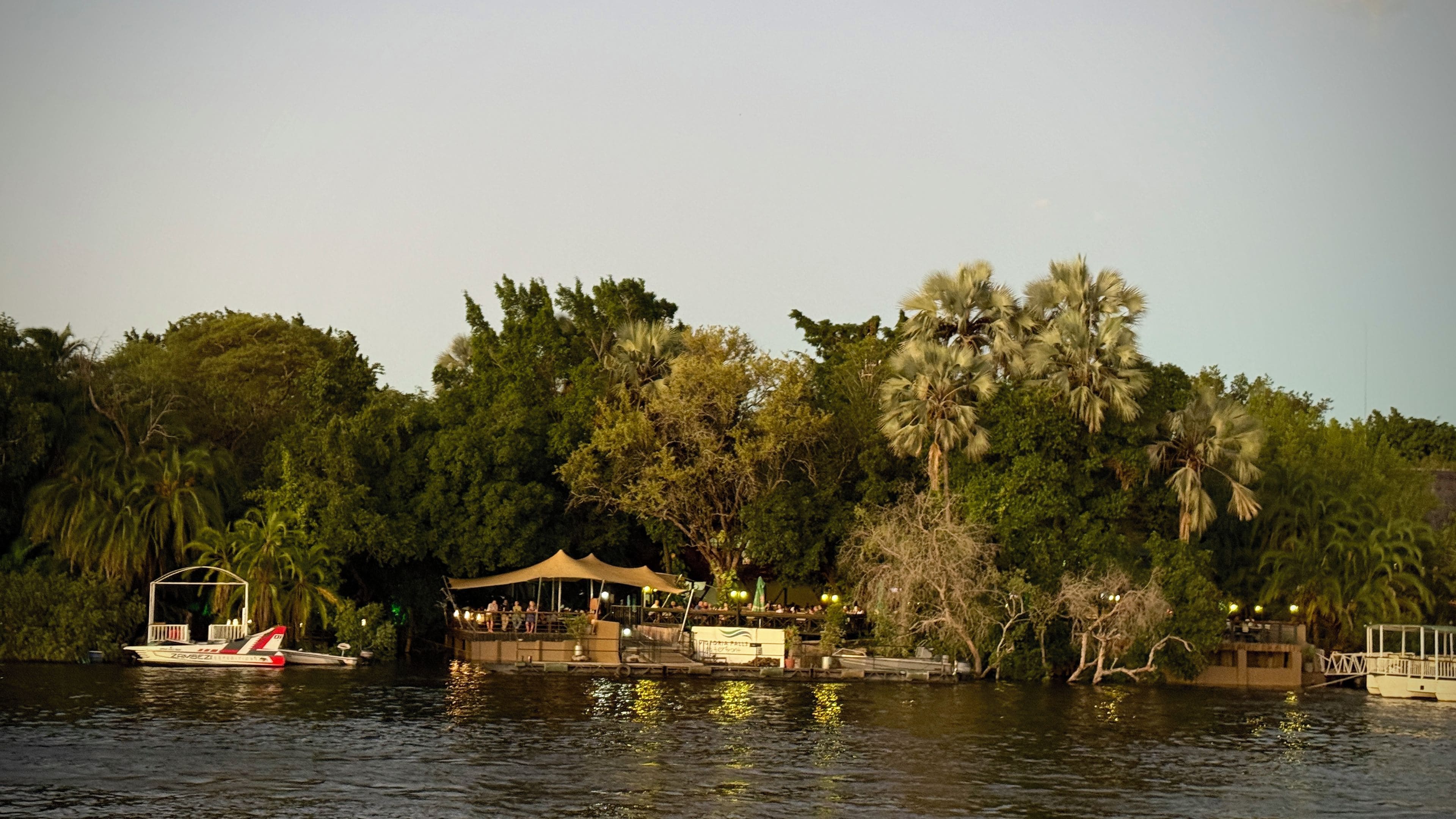 A riverside restaurant with outdoor seating, a boat docked nearby, and dense foliage in the background.