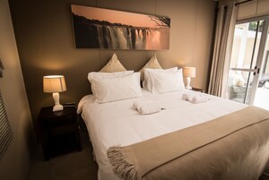 Una cama bien hecha con sábanas blancas y un edredón color beige, una mesita de noche con una lámpara, y un cuadro grande de una cascada en la pared.