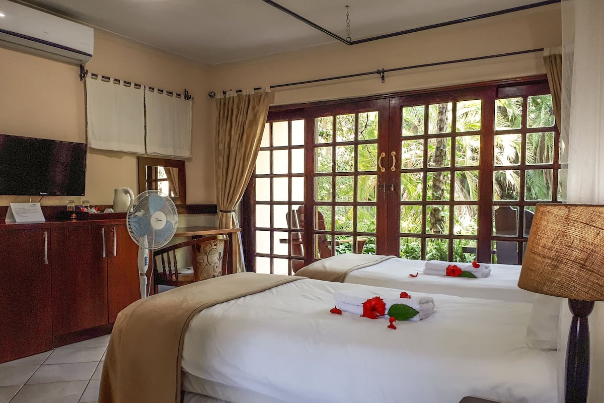 A hotel room with two beds, a TV, a fan, a desk with a computer, and a window with a view of greenery.
