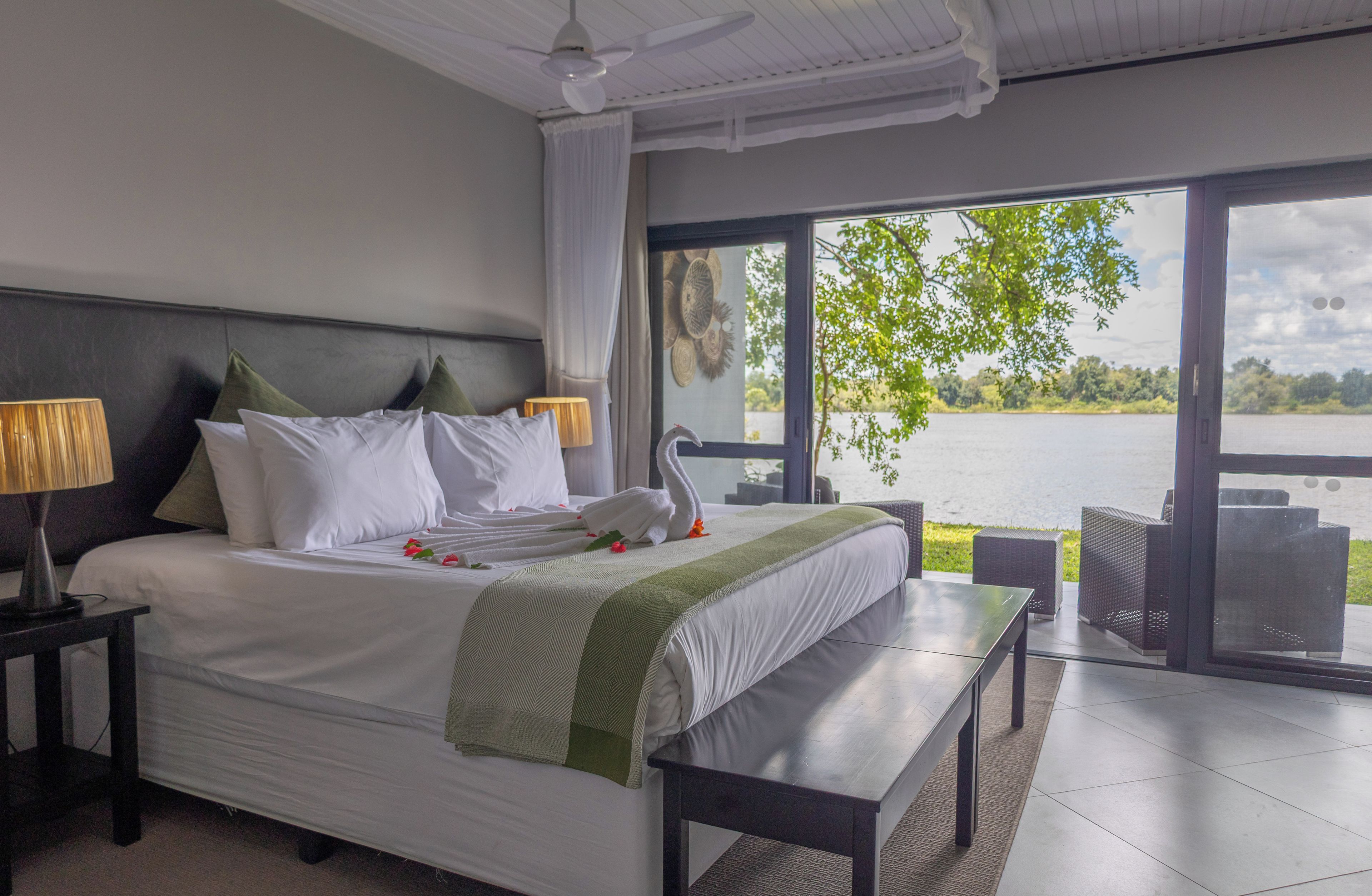 A hotel room with a large bed, two bedside lamps, a bench, and a view of a lake through large windows.
