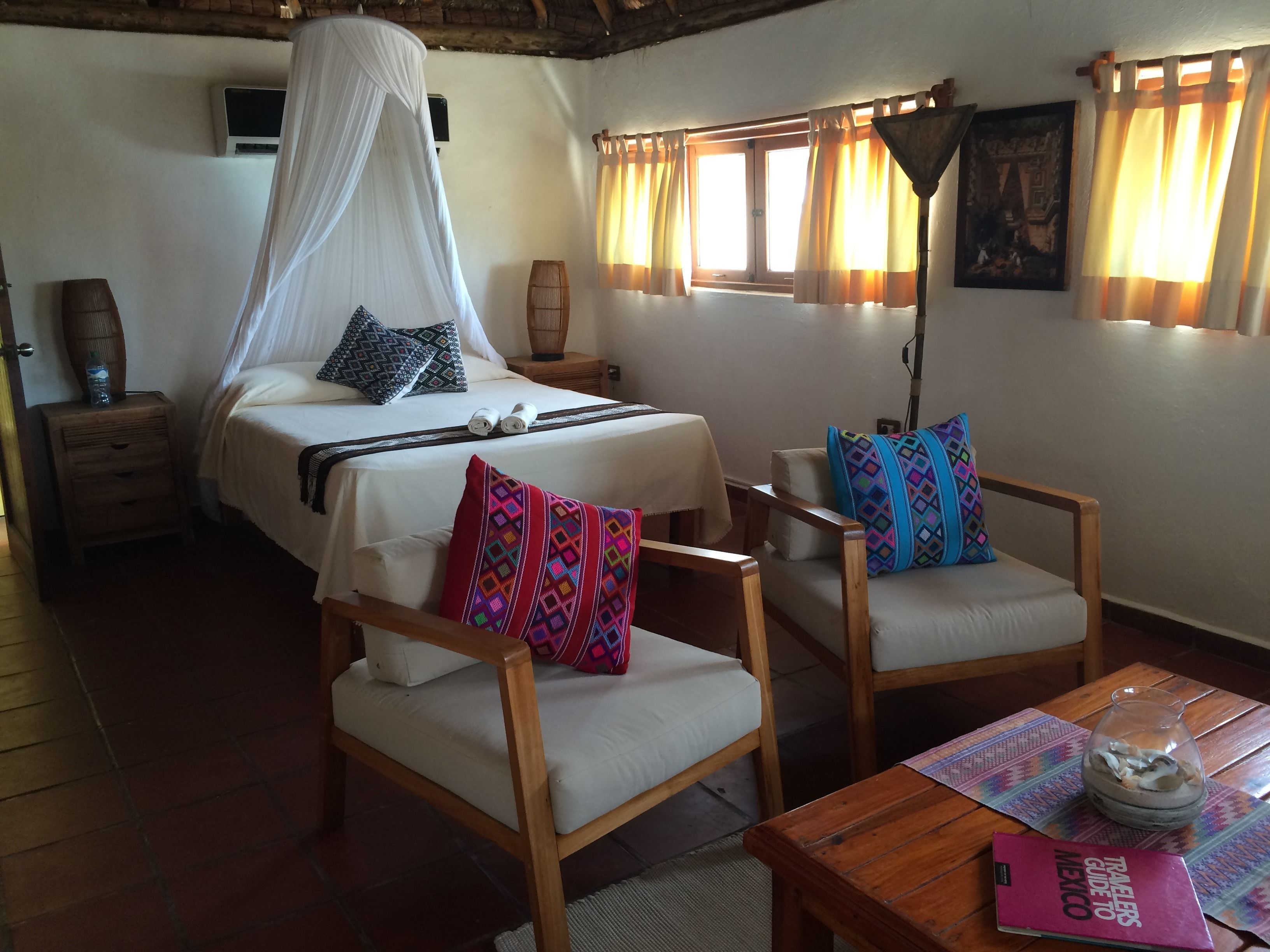 Quarto com cama, duas cadeiras de madeira, uma mesa e janela com cortinas.