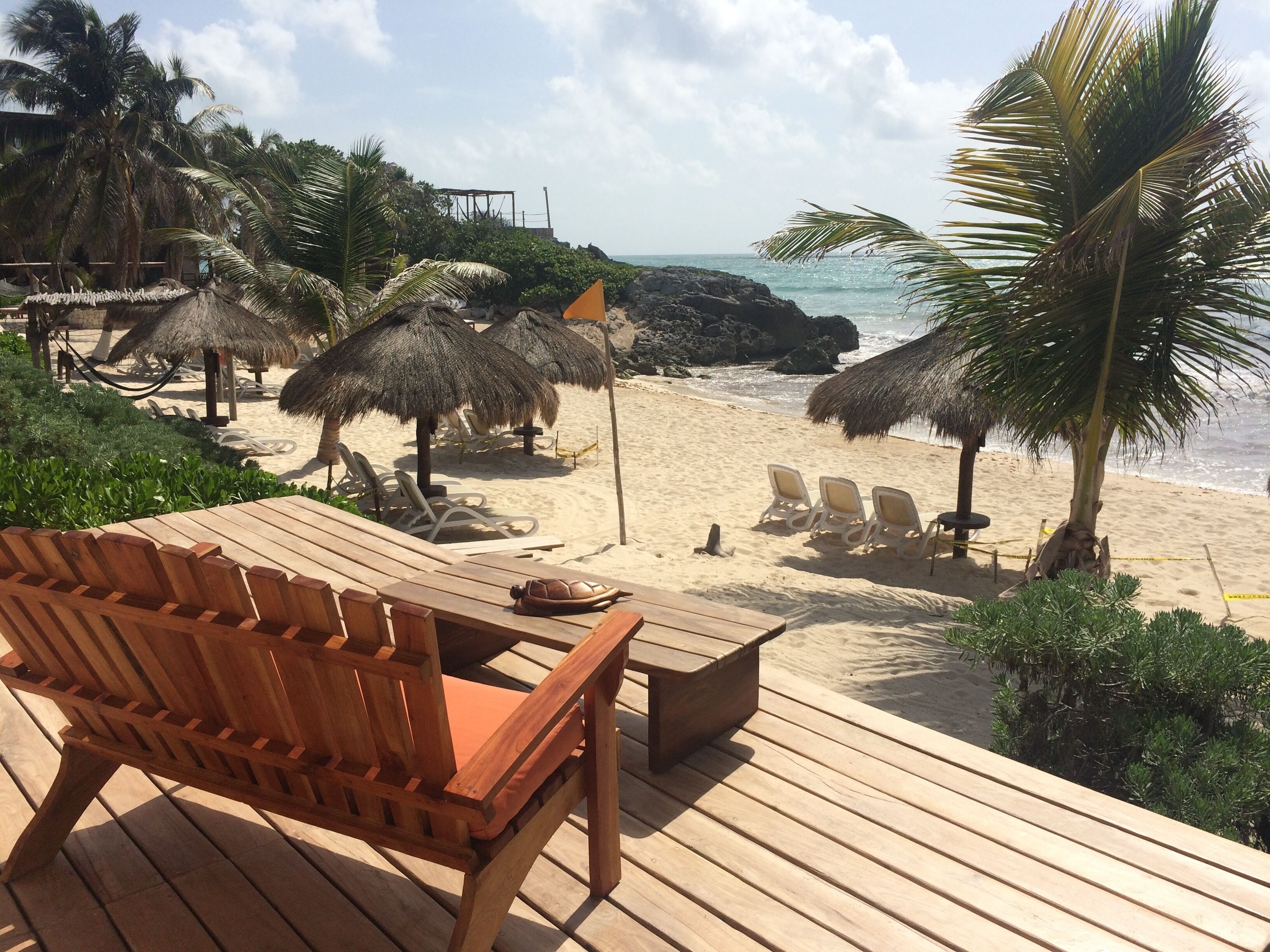 A wooden deck with lounge chairs and a table, overlooking a beach with thatched umbrellas and palm trees.