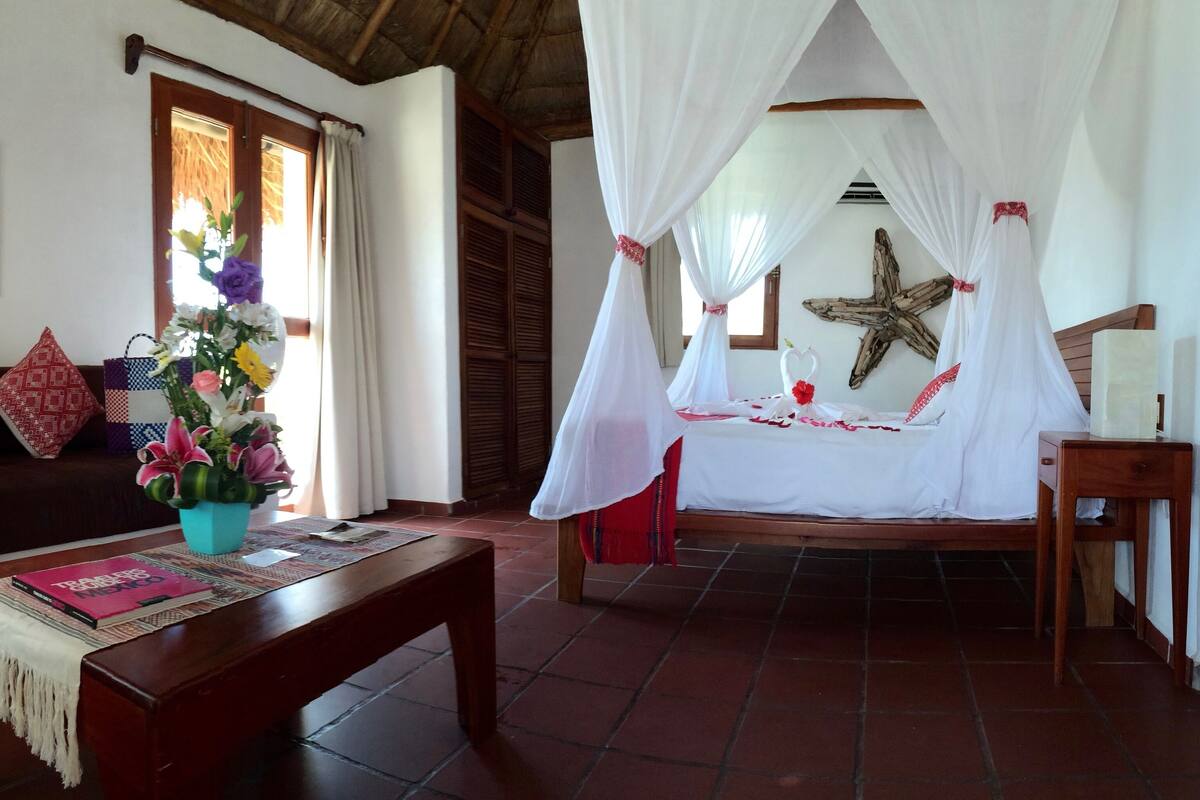 A room with a wooden bed under a white canopy, a wooden bench, a sofa with patterned cushions, a wooden table with a book, and a starfish decoration on the wall.