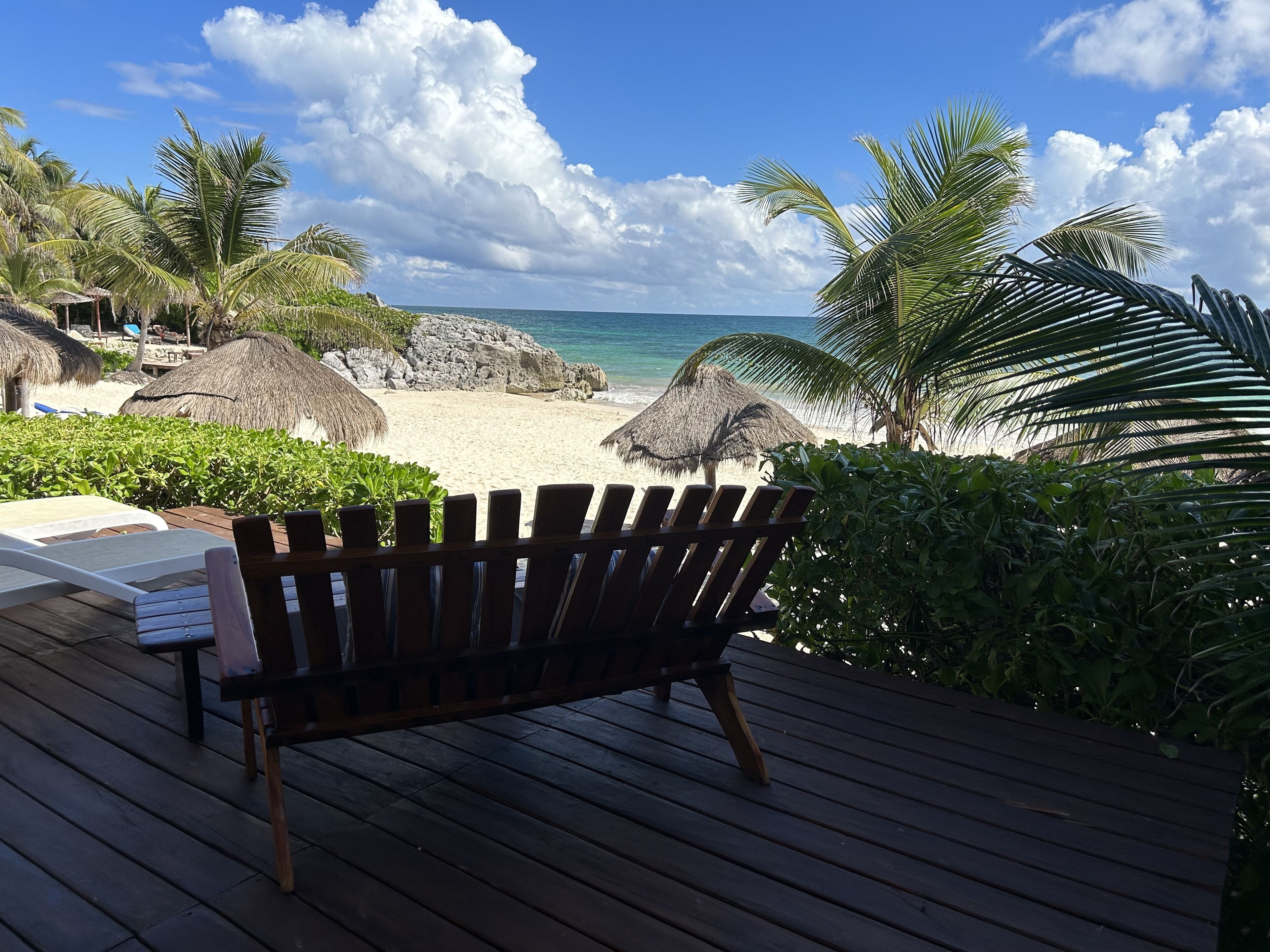 A wooden deck with a bench overlooking a beach with thatched umbrellas and palm trees.