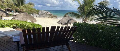 A wooden deck with a bench overlooking a beach with thatched umbrellas and palm trees.